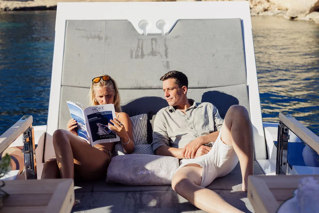 Paar liest das HÖKE-Yachting-Magazin auf der Sonnenliege einer Charter-Yacht vor Mallorca — sommerliches Lifestyle-Moment im Bademantel