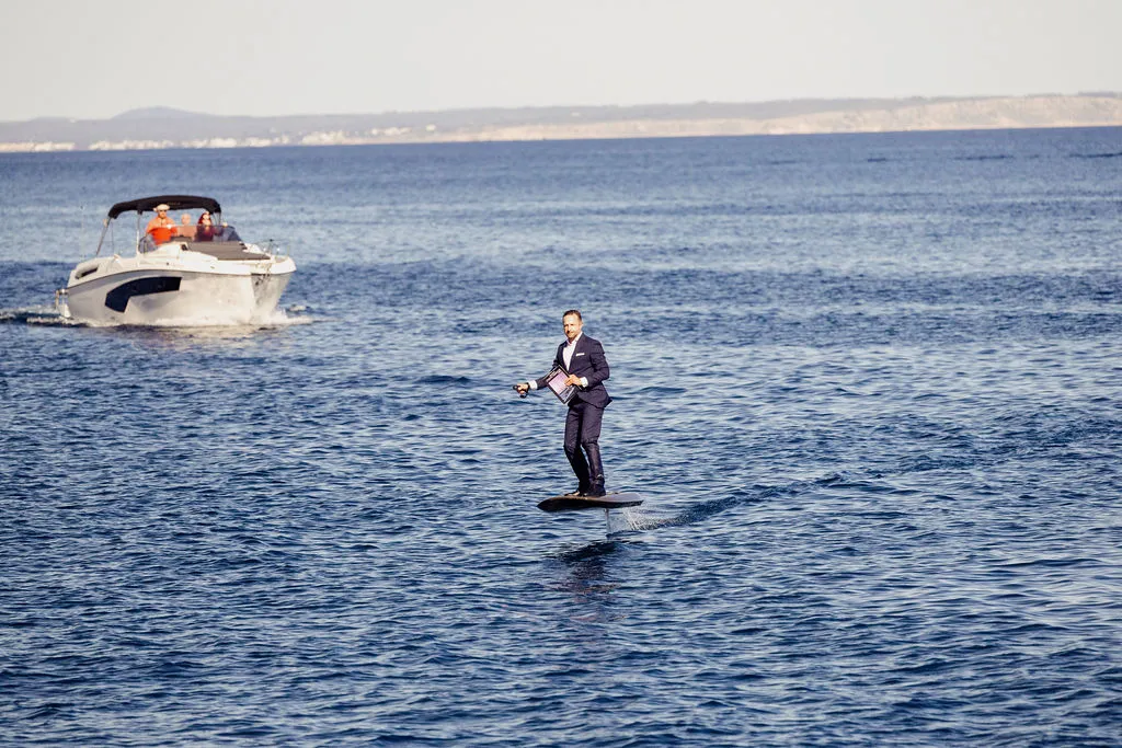 Julian Höke im Anzug auf einem E-Foil-Surfbrett vor Mallorca, das HÖKE-Yachting-Magazin in der Hand