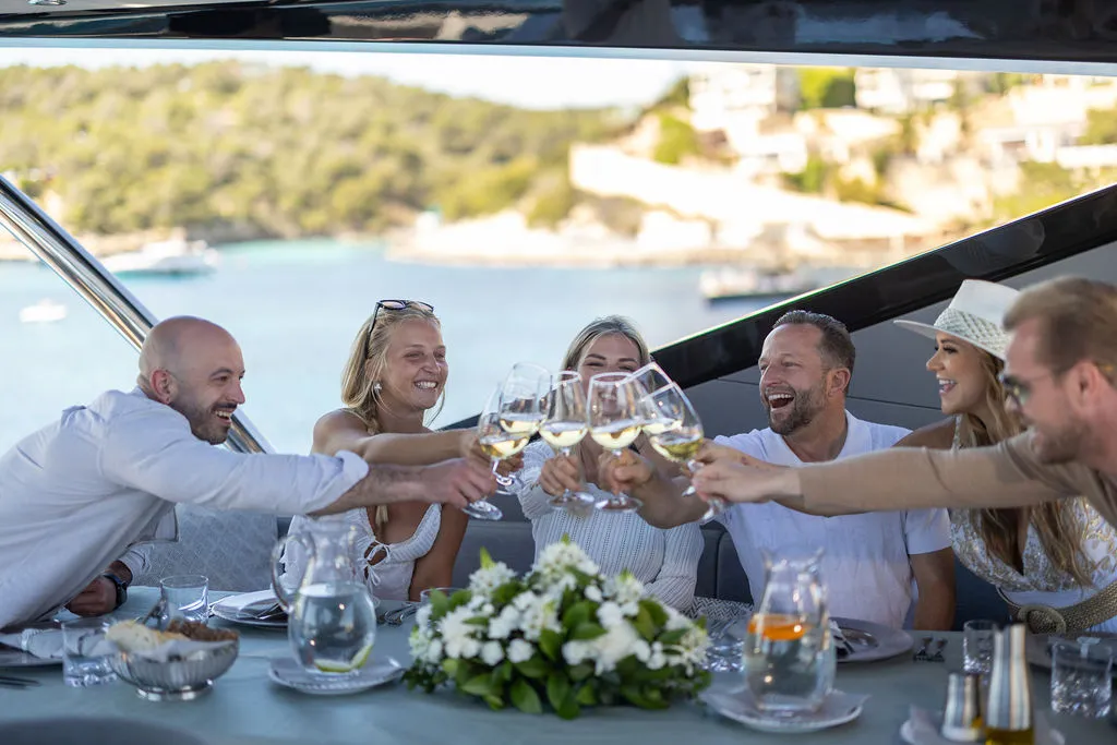 Charter-Gäste stoßen mit Weingläsern an Bord einer Yacht vor Mallorca an — sechs Personen feiern in einer ruhigen Bucht mit Hügelkulisse im warmen Tageslicht